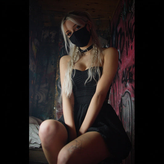 Woman in a black dress and face mask sitting against a graffiti-covered wall.