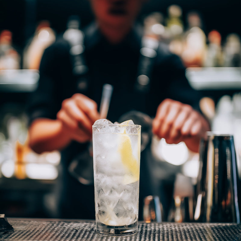 Barista preparing a cocktail with ice and lemon garnish at a bar.